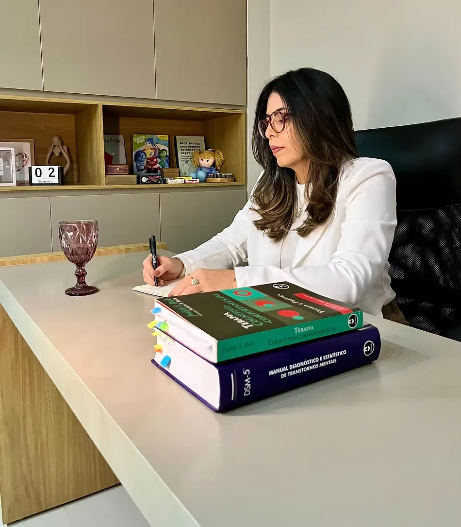Thaiza Leandro à mesa do consultório, escrevendo em um caderno ao lado de livros de TCC.