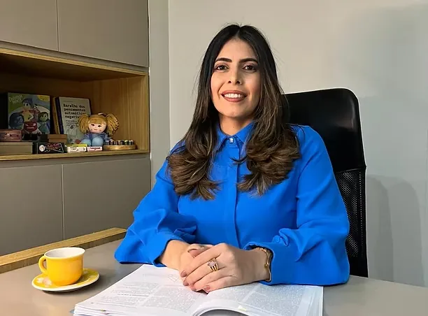 Thaiza Leandro de camisa azul, sorrindo, sentada à mesa do consultório.